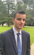 Iván Novegil, a young man in a dark suit and blue tie, photographed outdoors in a park with grass and trees in the background